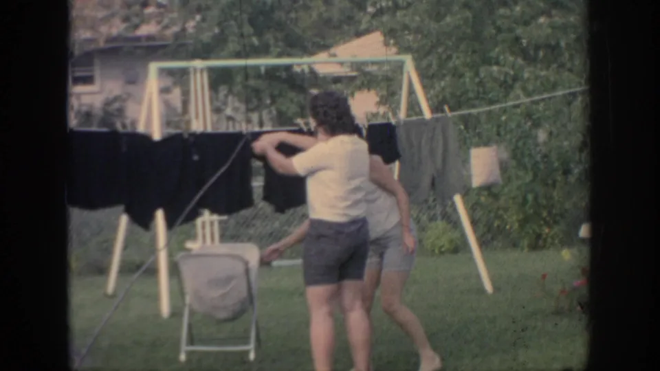 In the Oklahoma backyard, one woman adjusts dark laundry as another passes near the line.