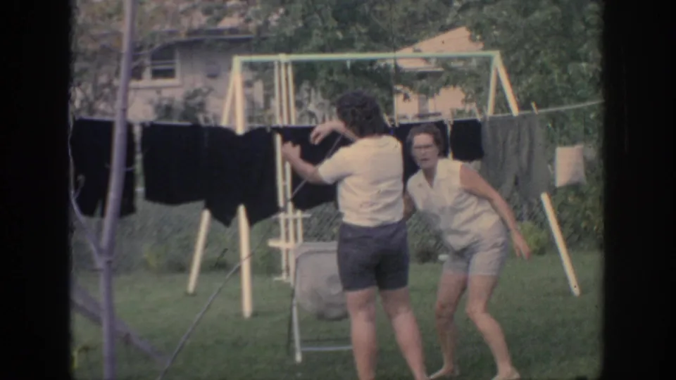 A woman continues hanging laundry as another turns toward the camera in the 1967 Oklahoma backyard.