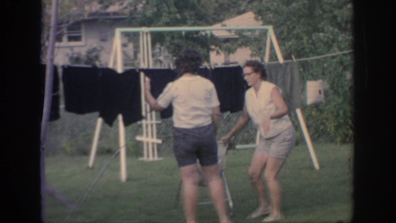 In 1967 Oklahoma home-movie footage, two women stand by dark laundry on a portable clothesline in a backyard with a swing set behind them.