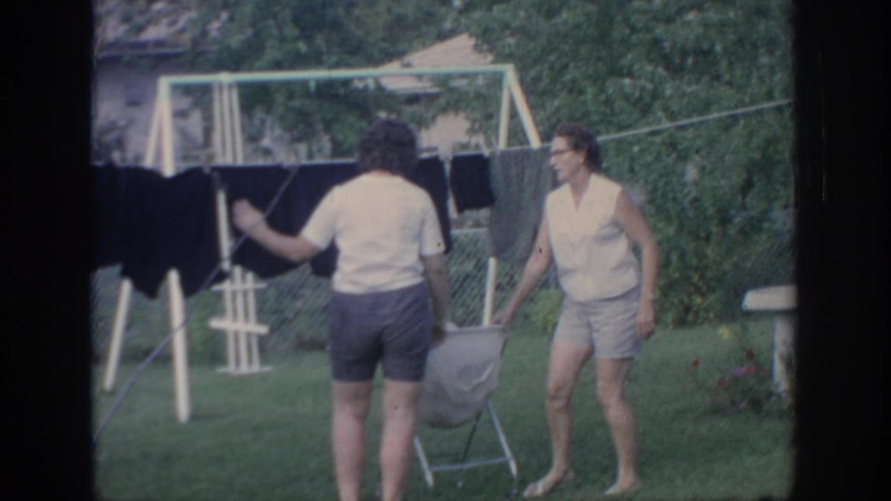 A woman in a white shirt handles laundry on a backyard clothesline as another woman in a sleeveless top stands near a folding basket.