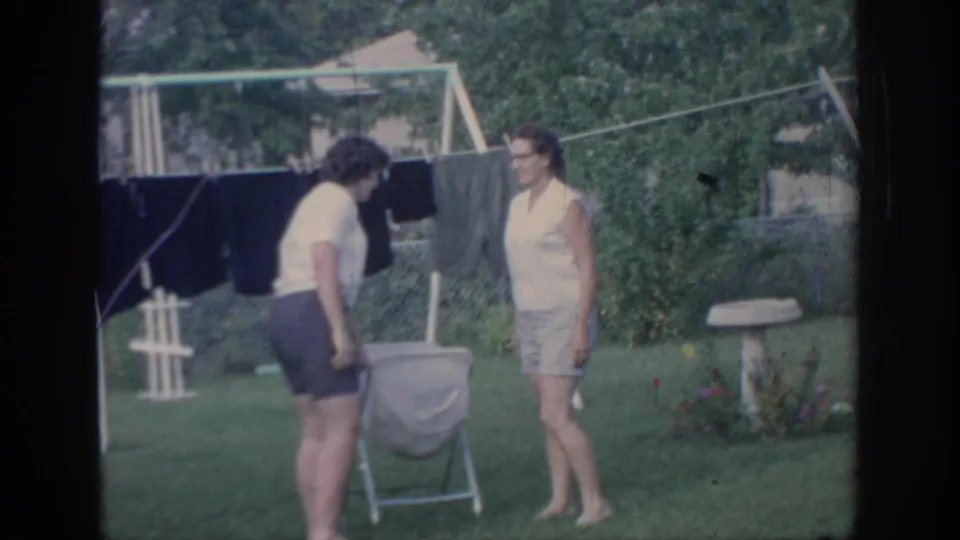 Two women face one another beside a folding basket and dark laundry, with backyard greenery and a bird bath visible nearby.