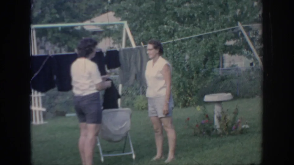 One woman holds a dark garment near the clothesline while another stands across from her in the 1967 Oklahoma backyard scene.