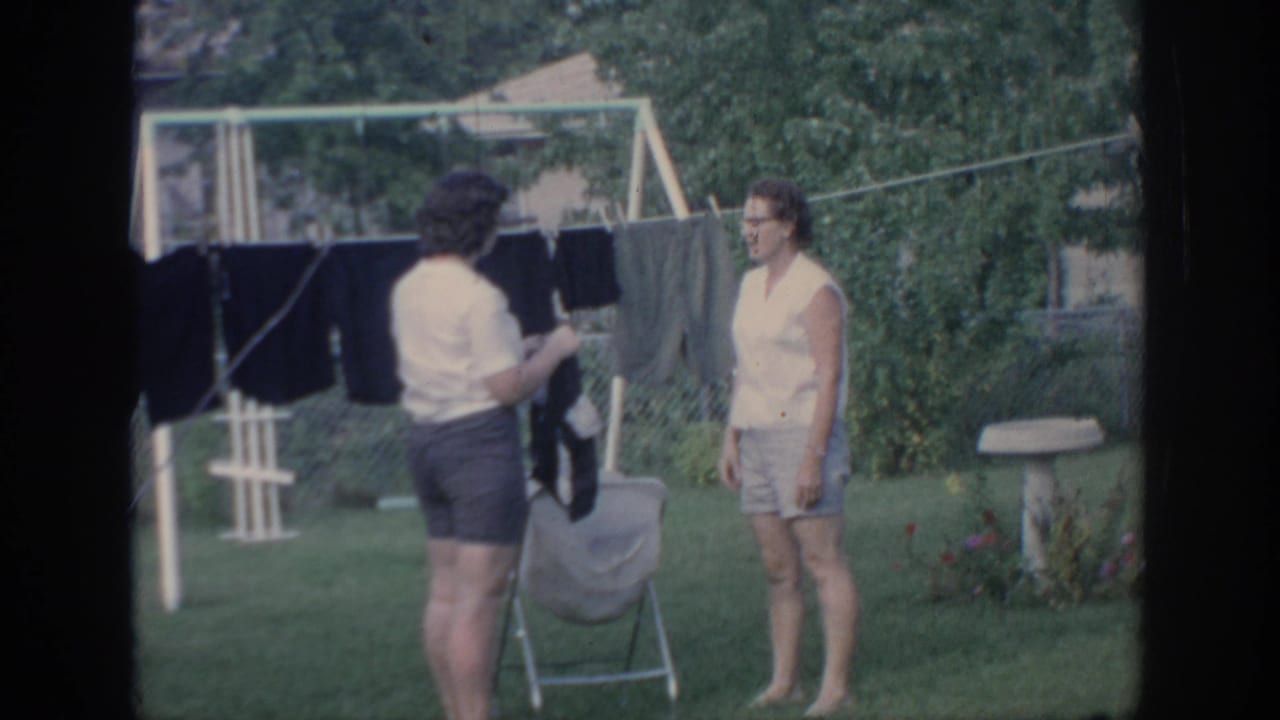 A woman holds laundry near a clothesline while another woman stands beside the yard.