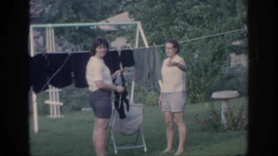 One woman faces the camera as the other holds laundry in the grassy Oklahoma backyard setting.