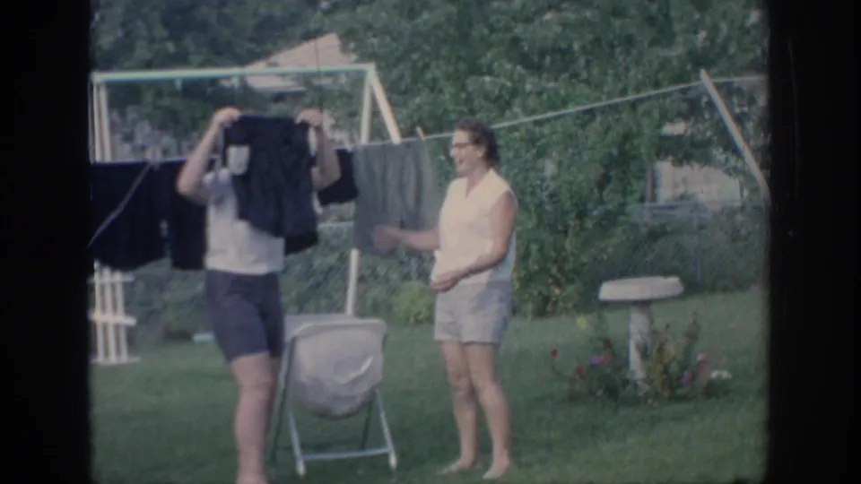 One woman raises a dark garment near the camera as another stands by the clothesline in the backyard.
