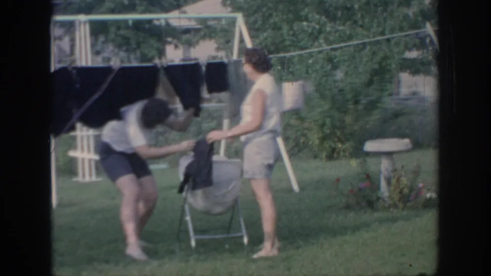 One woman bends toward garments as another stands beside a metal laundry cart in the 1967 Oklahoma backyard scene.