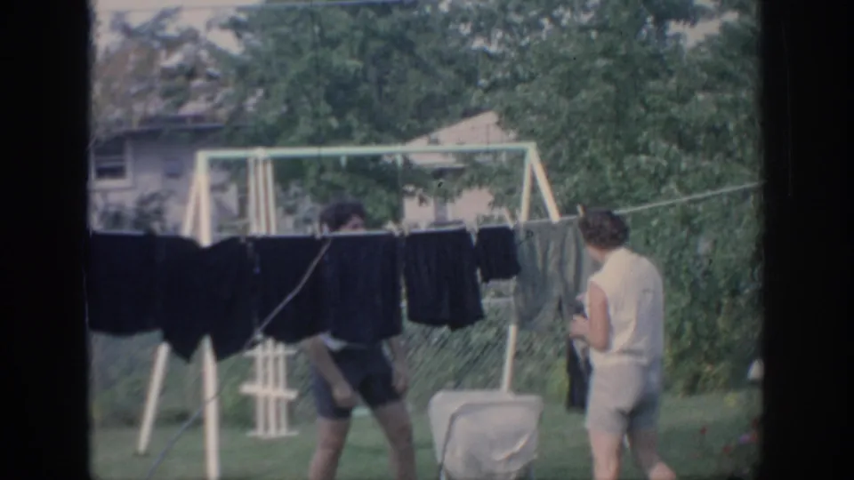 The women move behind dark garments on the clothesline, with the laundry cart and green backyard still visible.