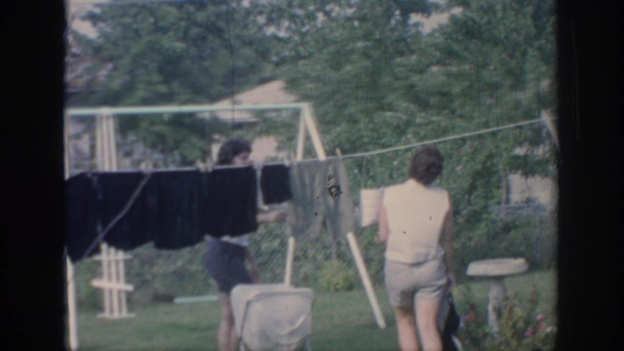 1967 Oklahoma home-movie frame showing two young women by a backyard clothesline with dark garments and a lawn chair nearby.