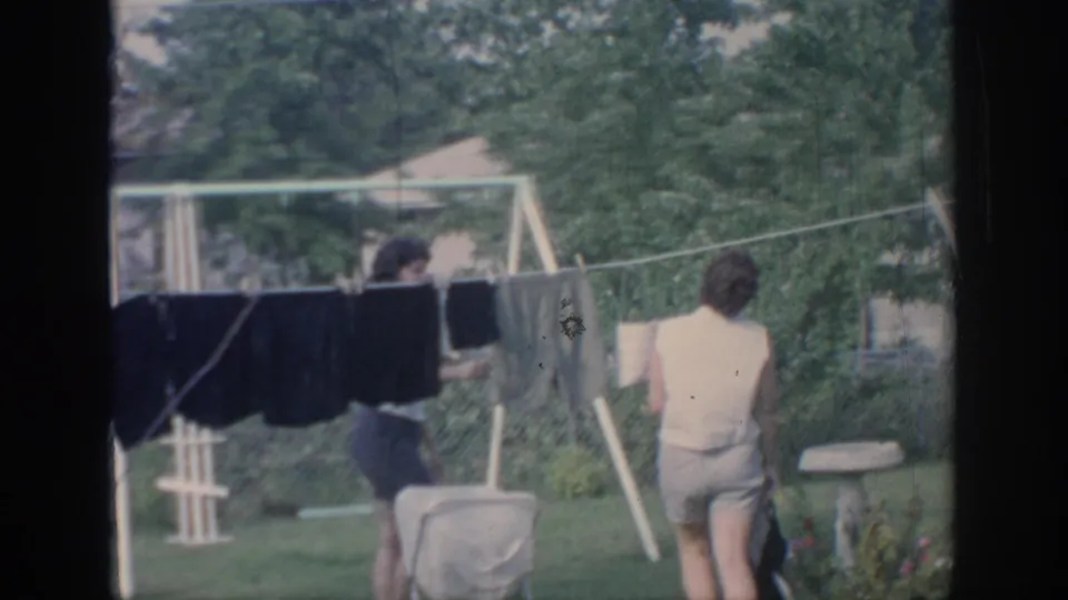 1967 Oklahoma home-movie frame showing two young women by a backyard clothesline with dark garments and a lawn chair nearby.
