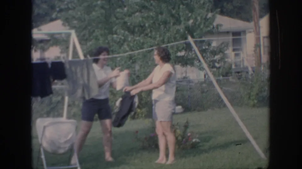 1967 Oklahoma home-movie frame of two young women playfully exchanging a garment beside a backyard clothesline.