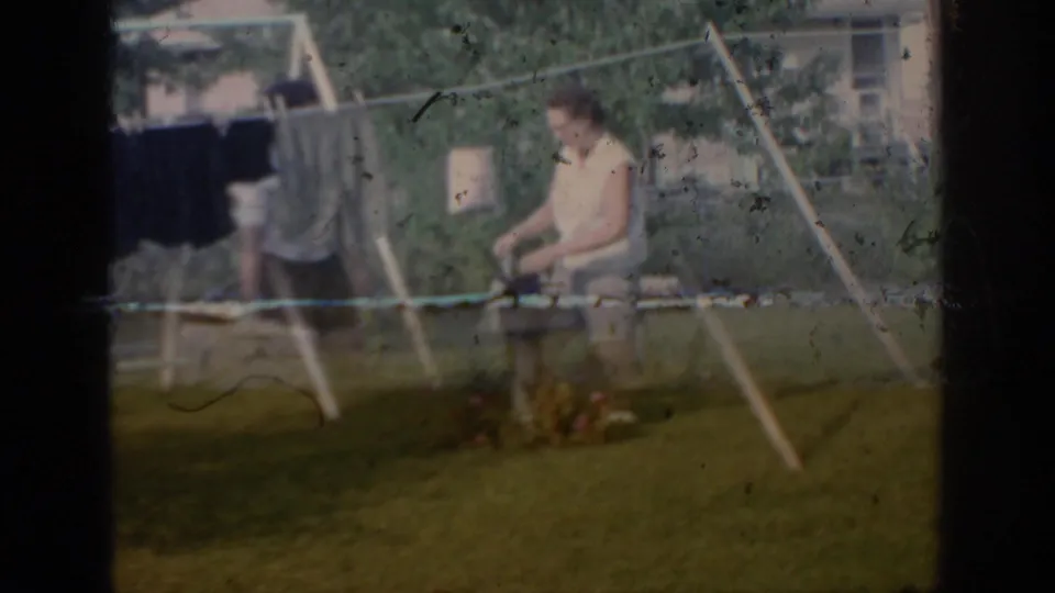 1967 Oklahoma home-movie frame showing a young woman bending by a backyard clothesline with garments in the foreground.