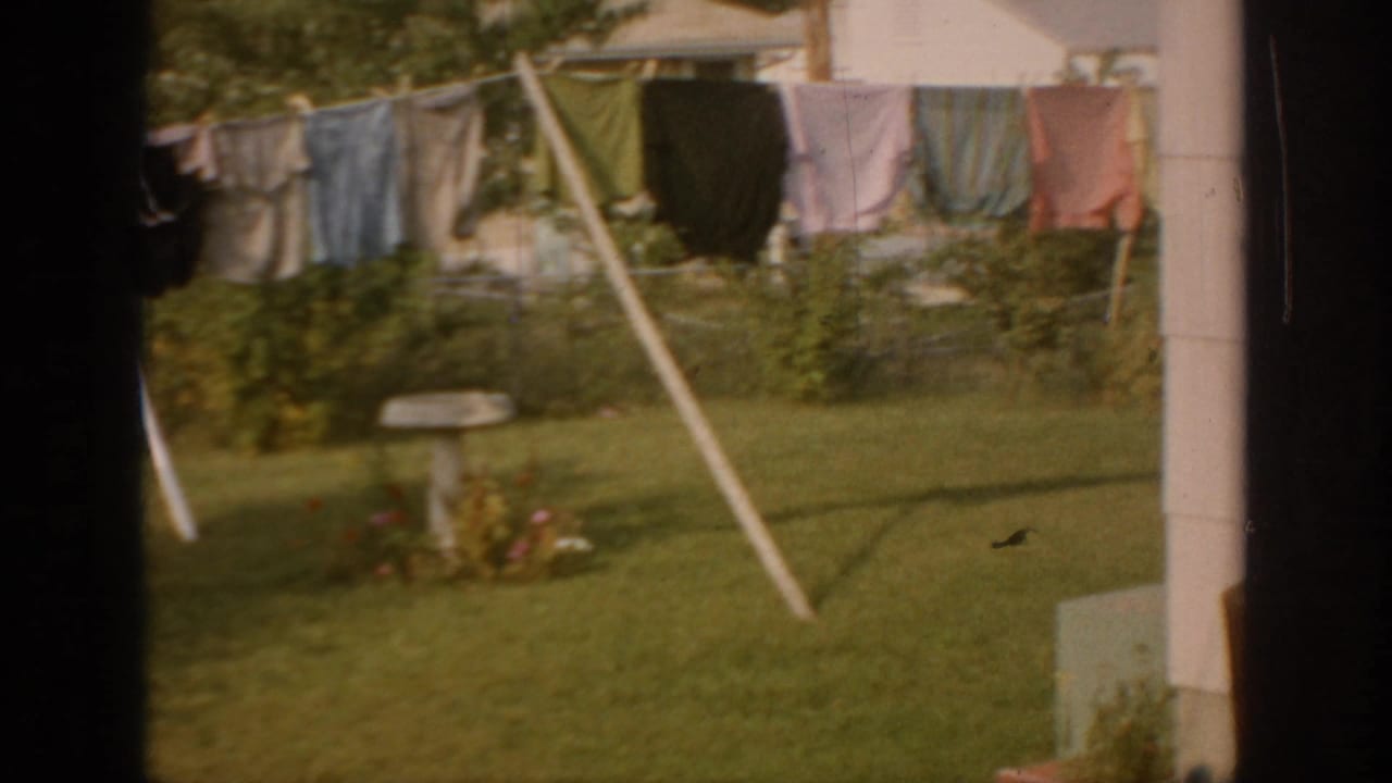 1967 Oklahoma 8mm frame showing colorful laundry on a backyard clothesline, green lawn, shrubs, and a small garden feature.