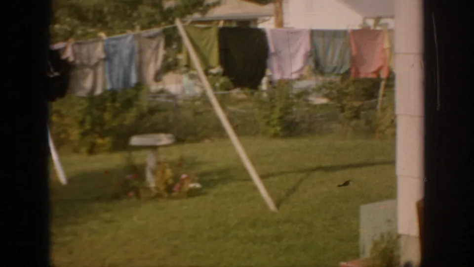 1967 Oklahoma 8mm frame showing colorful laundry on a backyard clothesline, green lawn, shrubs, and a small garden feature.