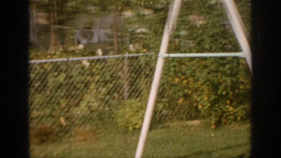 1967 Oklahoma backyard frame with chain-link fence, dense shrubs, lawn, and an angled white support pole in warm 8mm color.