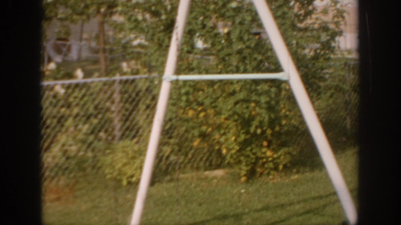 1967 Oklahoma backyard view with green lawn, shrubs, chain-link fence, house exterior, and a white A-frame yard structure.