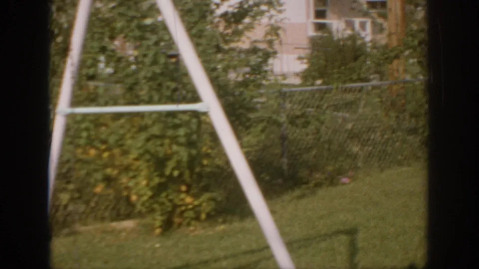 1967 Oklahoma home-movie frame showing lawn, shrubs, chain-link fence, nearby house details, and white A-frame supports.