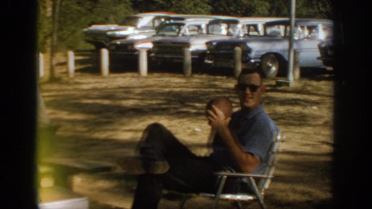 1967 Oklahoma home-movie frame showing a man in sunglasses reclining in a folding chair with a football near late-1960s cars.