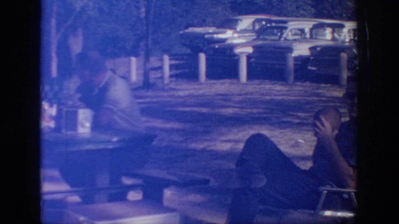 Blue-tinted 8mm frame from the Oklahoma picnic showing a seated man with a football and parked cars in the background.