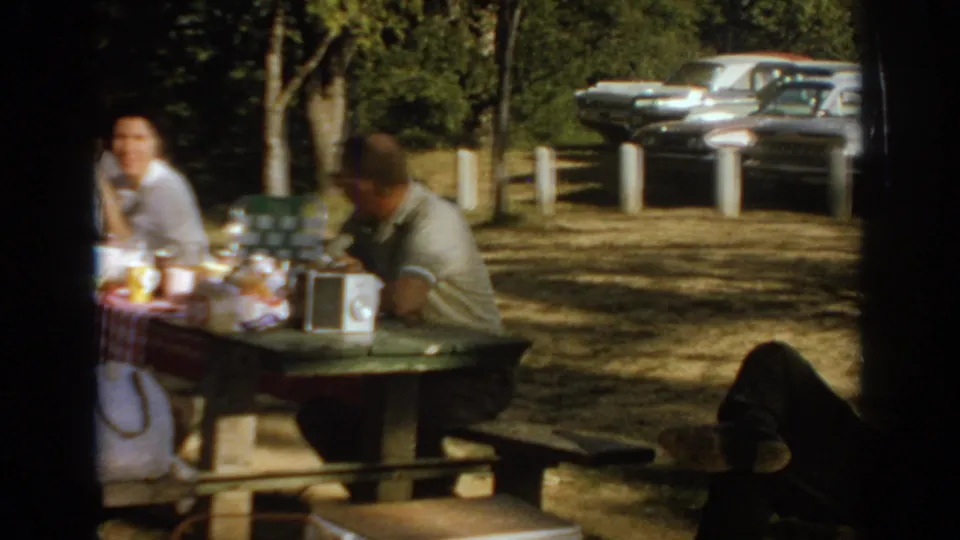 1967 Oklahoma picnic frame with adults at a table, a checkered tablecloth, picnic items, portable radio, and parked cars behind them.