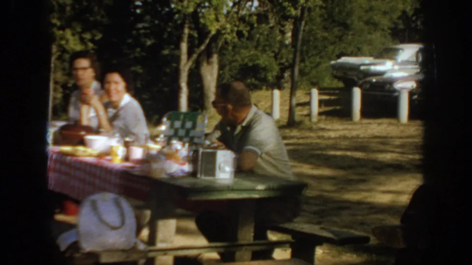 1967 Oklahoma home-movie frame showing three adults at a picnic table with a radio, picnic items, trees, and late-1960s cars nearby.