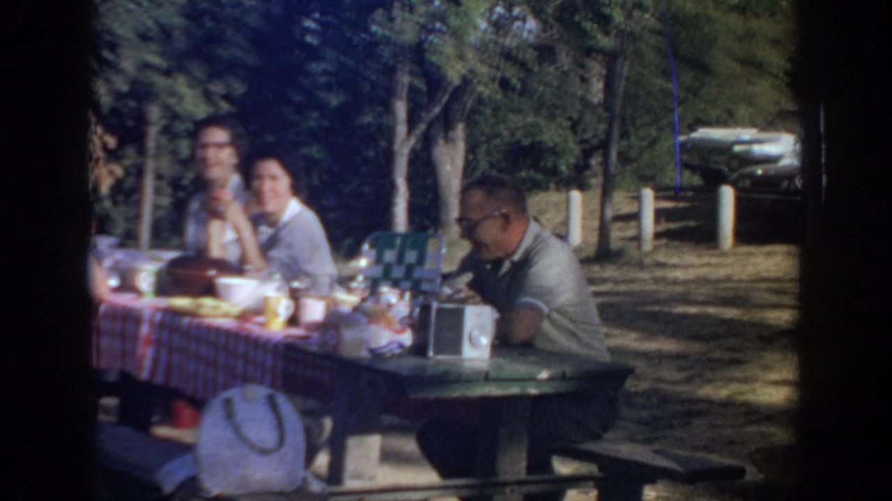 1967 Oklahoma home-movie frame of adults at a red checkered picnic table with food, cups, a portable radio, trees, and a parked car beyond.