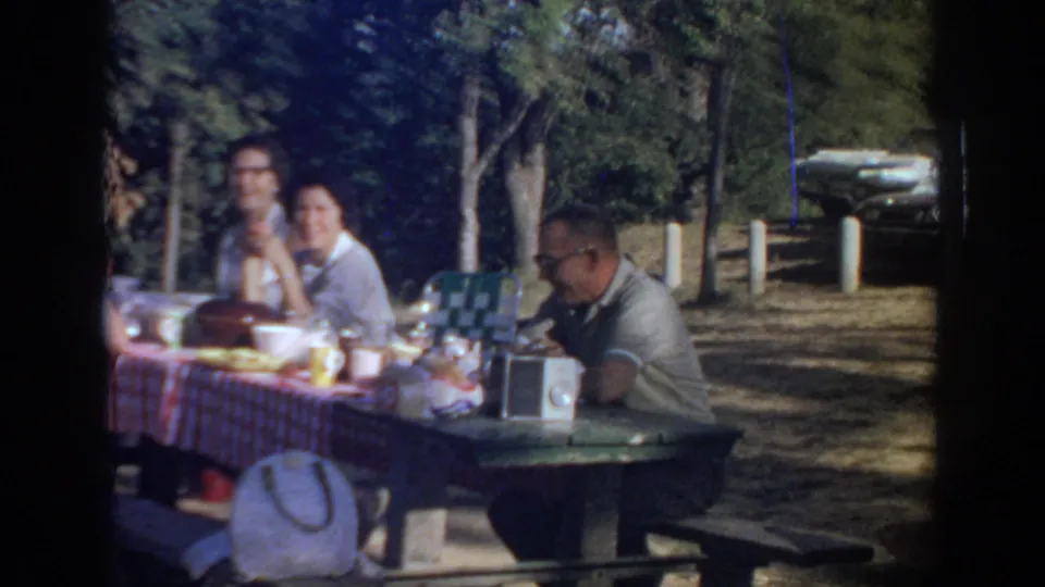 1967 Oklahoma home-movie frame of adults at a red checkered picnic table with food, cups, a portable radio, trees, and a parked car beyond.