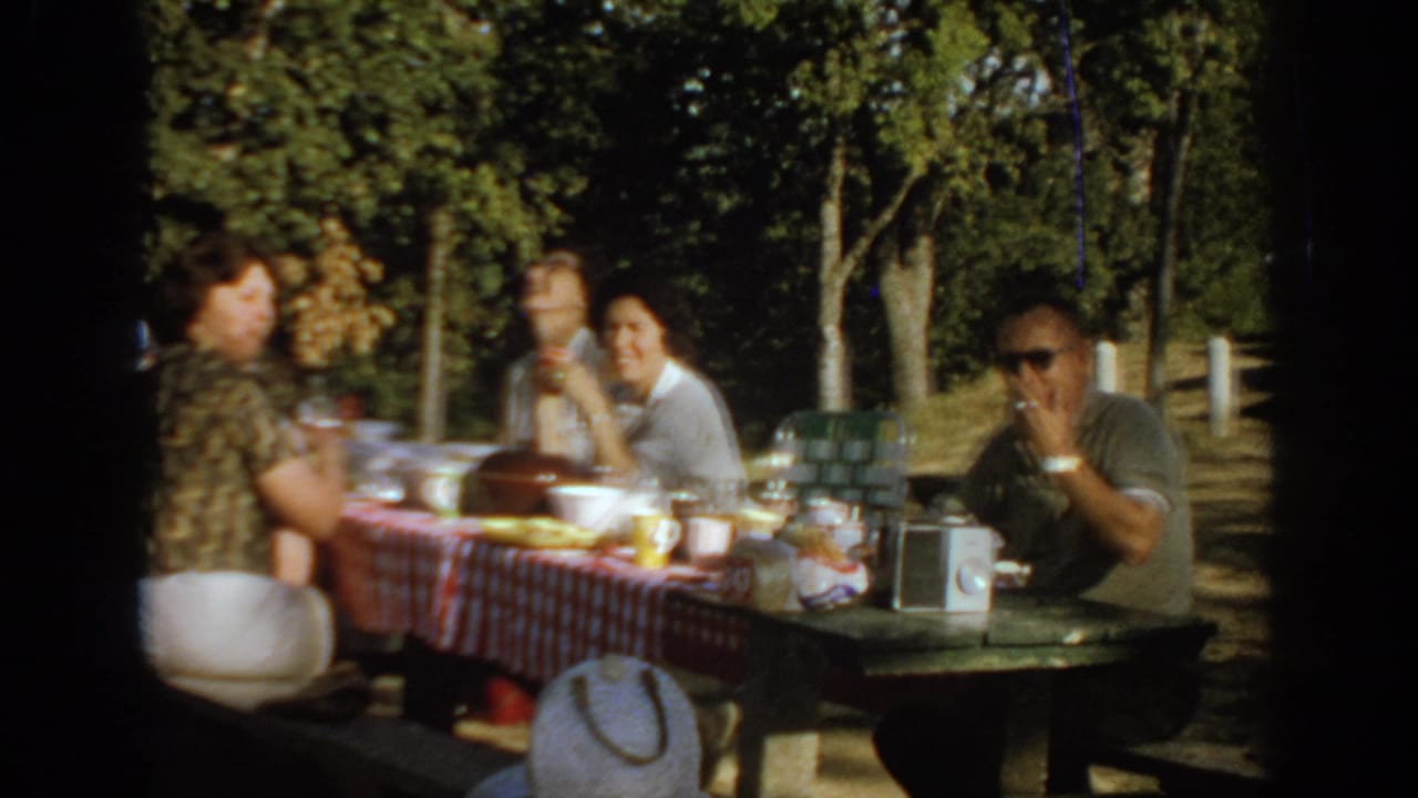 1967 Oklahoma picnic frame showing adults around a table, food and cups on the cloth, and a man in sunglasses smoking near the camera.