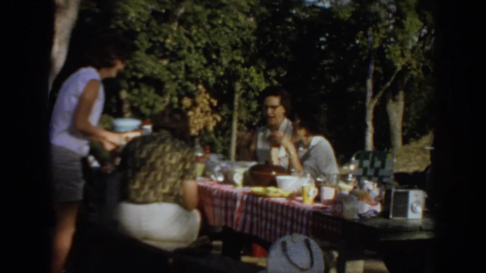 1967 Oklahoma home-movie frame with a standing person serving or reaching near a picnic table where adults sit with food, drinks, and a radio.