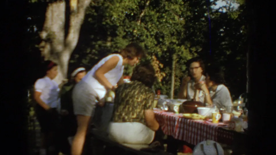 1967 Oklahoma picnic frame with people gathered under trees, one person bending near the table, and food, cups, and tableware spread out.