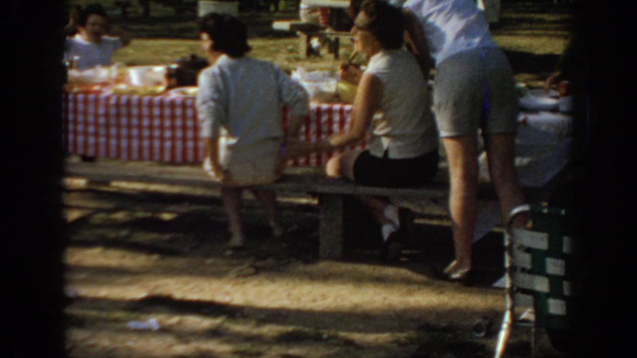 Women sit and stand near a picnic table with a red-and-white checkered tablecloth.
