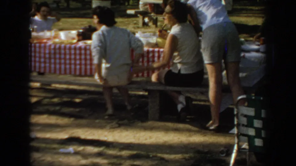 In a 1967 Oklahoma park scene, women gather around a picnic table covered with a red-and-white checkered tablecloth under tree shade.