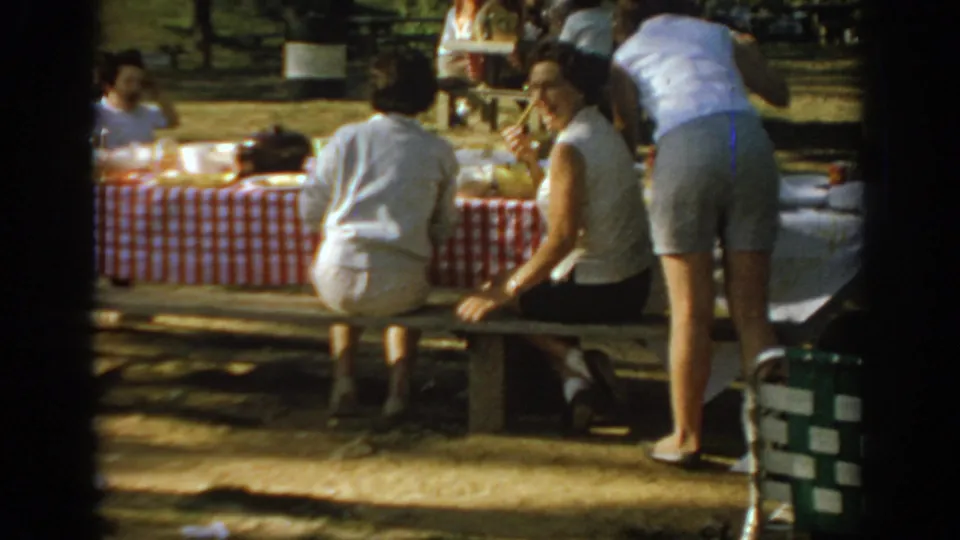 Women in 1960s casual clothing sit and stand beside a checkered picnic table in an Oklahoma park, with a green folding chair nearby.