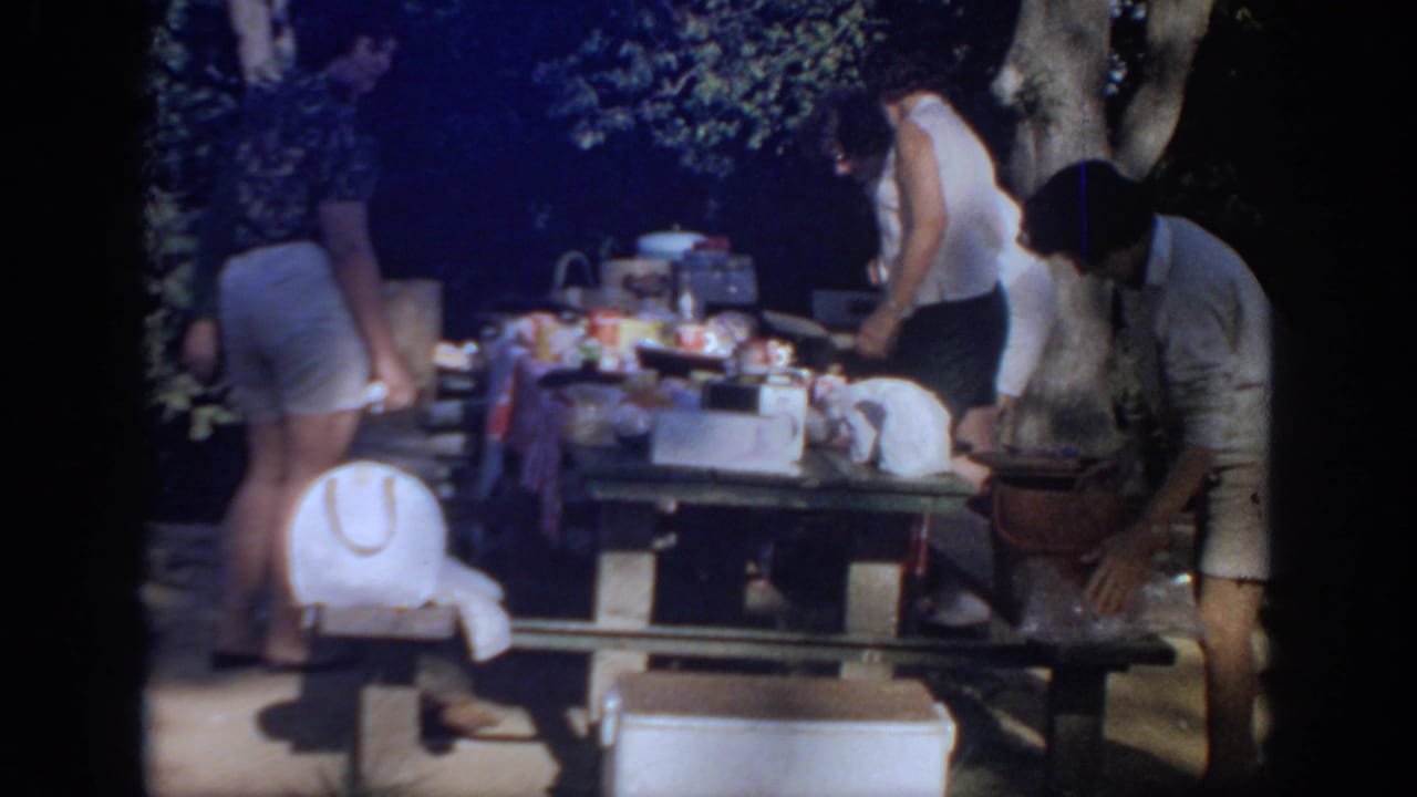 1967 Oklahoma home-movie frame of women preparing food at a shaded outdoor picnic table with a white cooler in front.