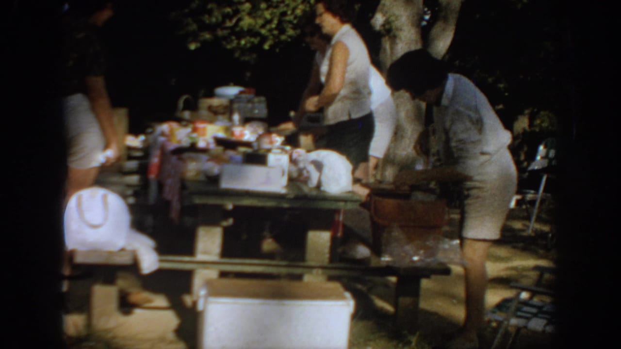 Women prepare a picnic table under trees, with a cooler and supplies in the foreground.