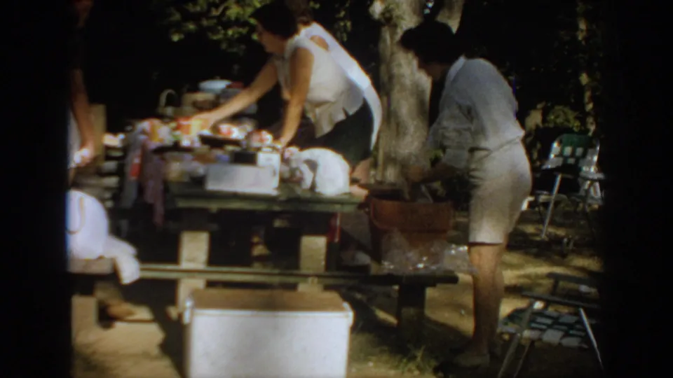 Women in casual 1960s summer clothing prepare an outdoor cookout table in Oklahoma home-movie footage.