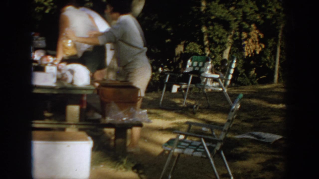 1967 Oklahoma home-movie frame showing adults beside a picnic table with food, folding chairs, a cooler, and trees in a shaded campsite.