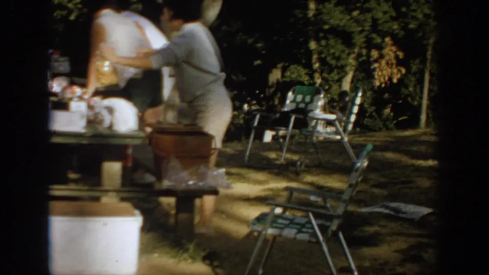 1967 Oklahoma home-movie frame showing adults beside a picnic table with food, folding chairs, a cooler, and trees in a shaded campsite.