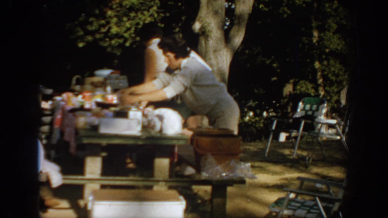 1967 Oklahoma picnic scene with an adult reaching over table supplies, another adult nearby, folding chairs, cooler, and trees visible.