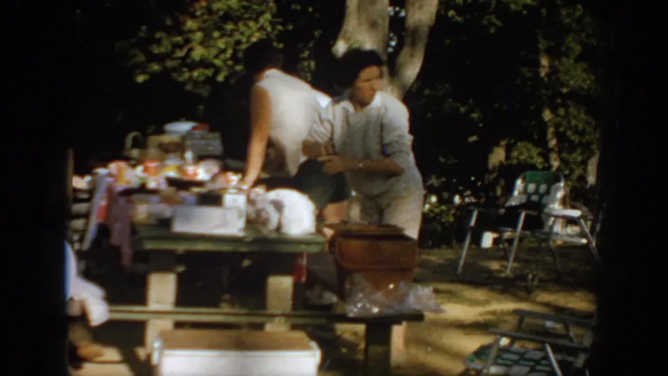 1967 Oklahoma frame showing two adults beside a picnic table at a wooded campsite, with food items, chairs, and a cooler visible.