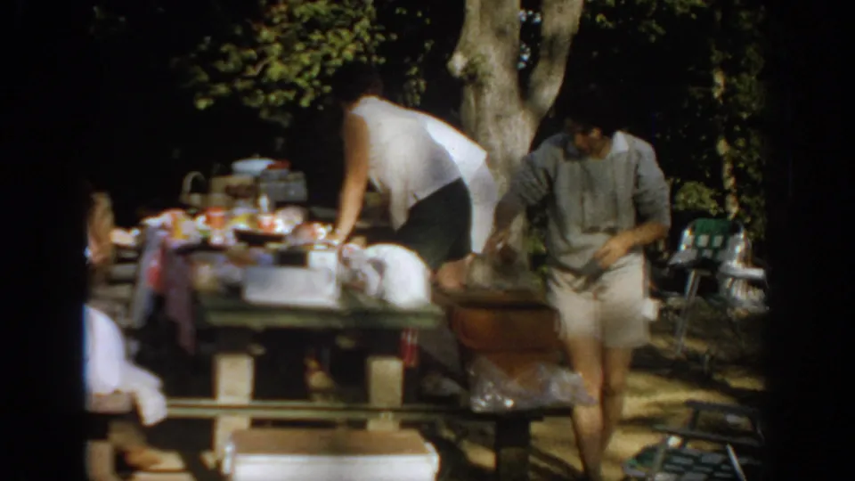1967 Oklahoma home-movie frame with one adult seated and another standing by a picnic table in a shaded wooded campsite.