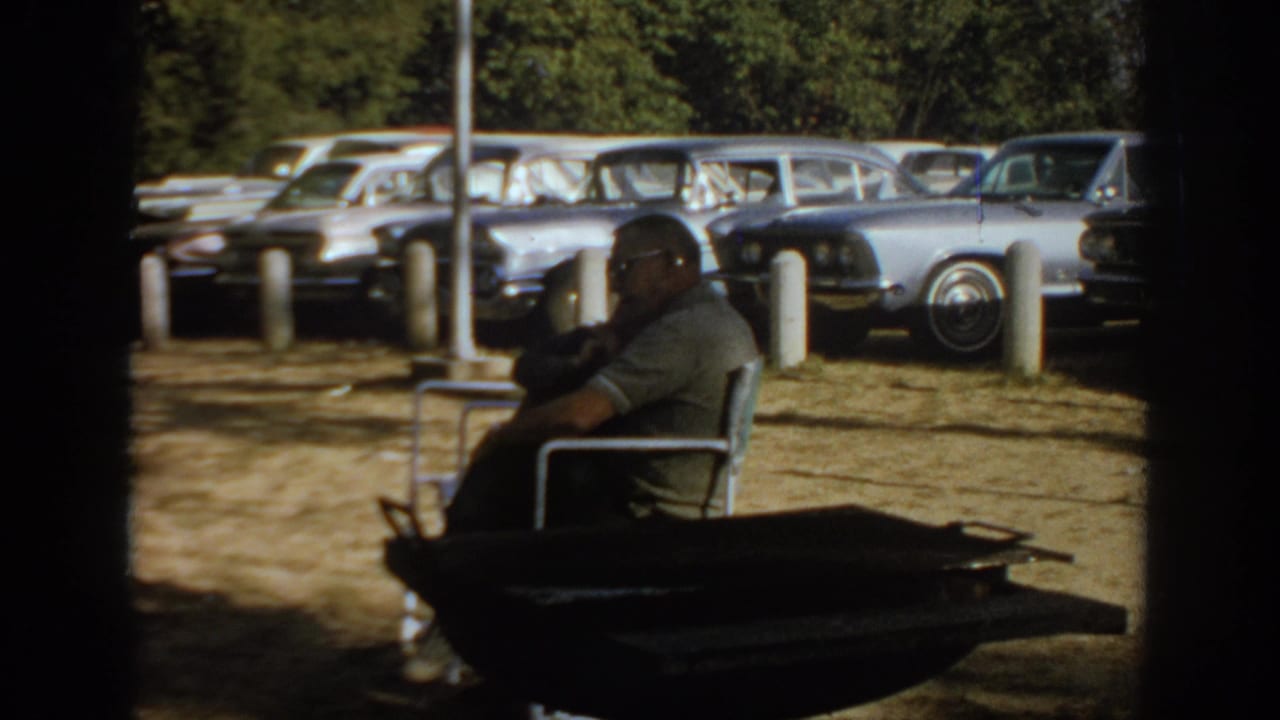 1967 Oklahoma home-movie frame showing a man in sunglasses seated in a metal chair with 1960s cars parked behind him.