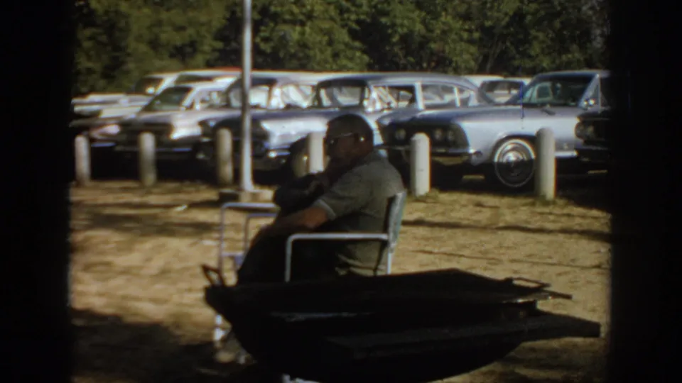 1967 Oklahoma home-movie frame showing a man in sunglasses seated in a metal chair with 1960s cars parked behind him.