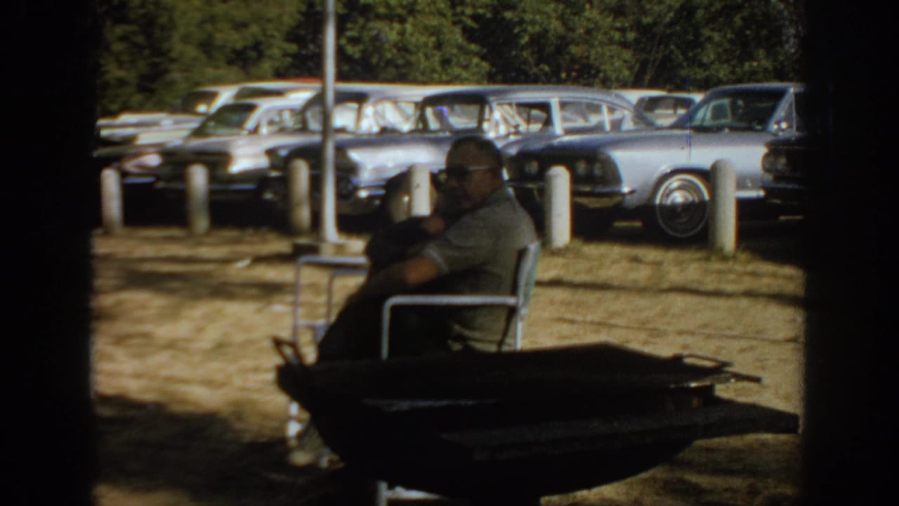 Seated man beside a dark foreground object with 1960s cars behind him.