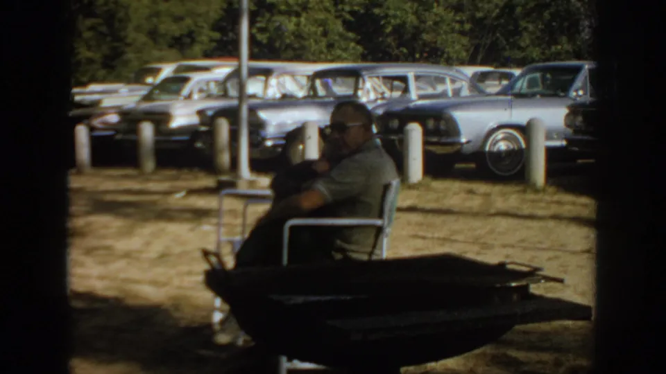 A man in a light shirt and sunglasses sits outdoors in Oklahoma, with classic cars and trees in the background.