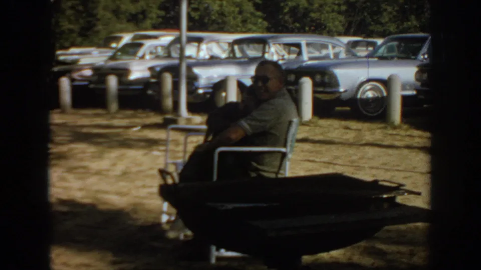Home-movie frame of a seated man at a park in 1967 Oklahoma, with a row of mid-century cars in the background.