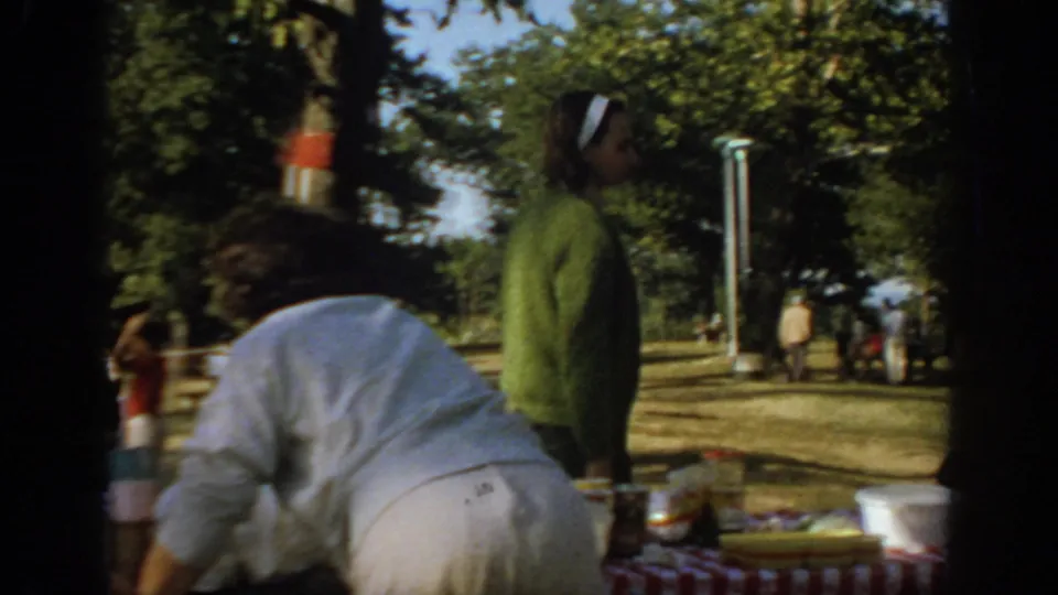 1967 Oklahoma picnic scene with adults near a checkered tablecloth, trees, and park fixtures in the background.