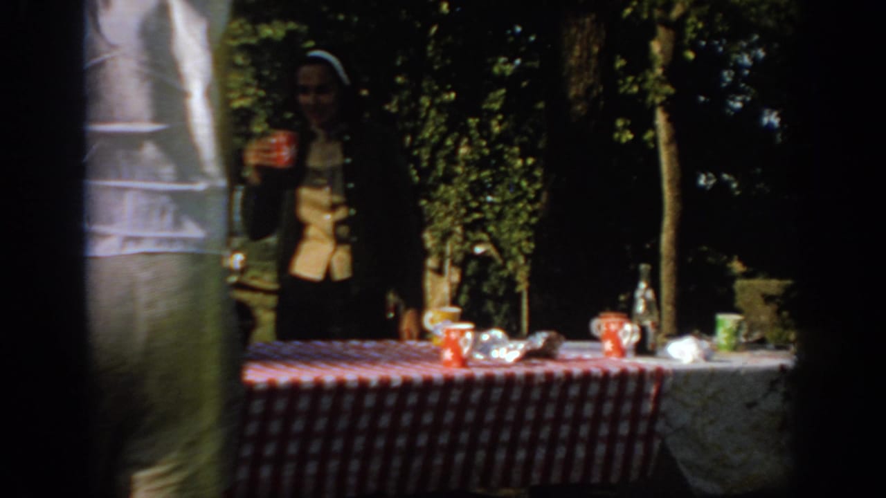 1967 Oklahoma 8mm frame of a woman in a headscarf holding a red cup behind a checkered picnic table in a shaded park.