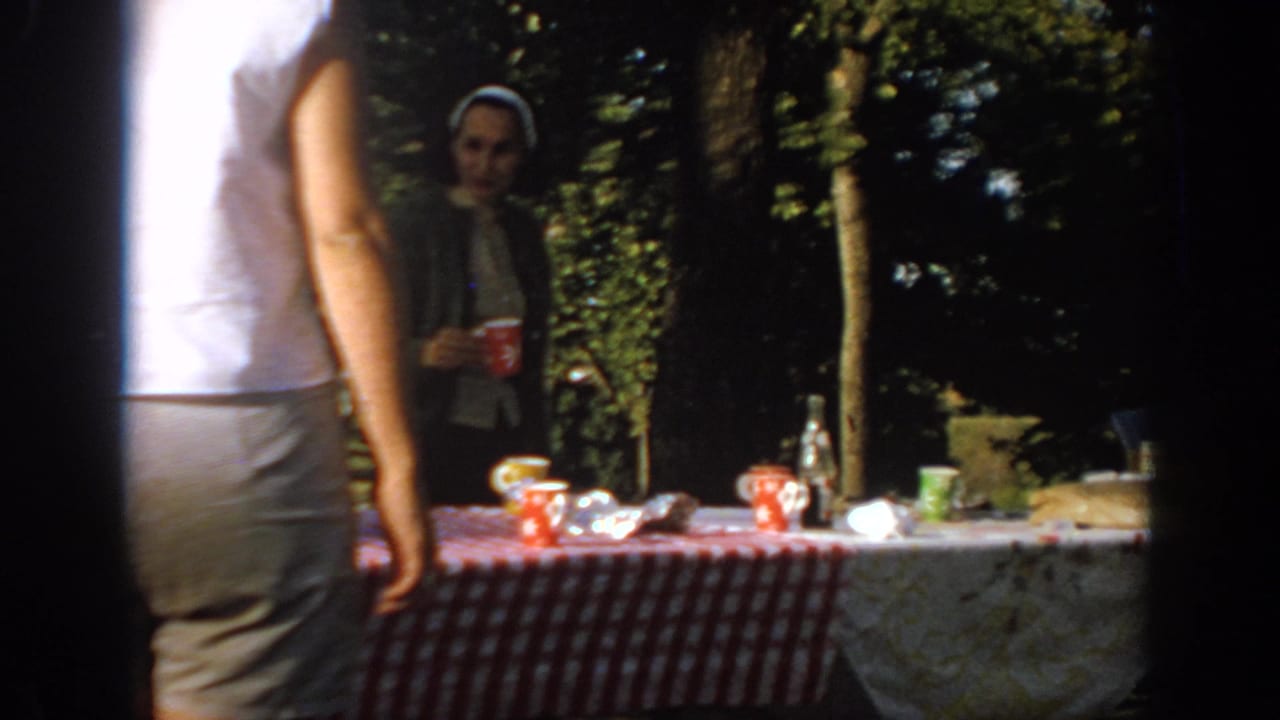 1967 Oklahoma home-movie frame showing an adult in a white sleeveless shirt near a park picnic table with cups and bottles.