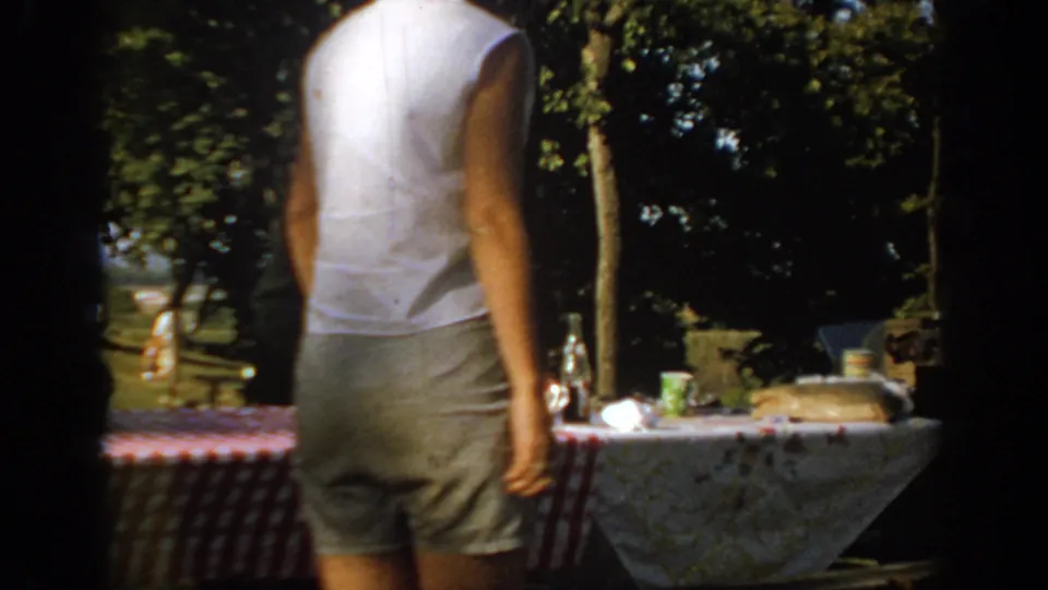 1967 Oklahoma 8mm frame of an adult seen from behind beside a checkered picnic table in a sun-dappled park.