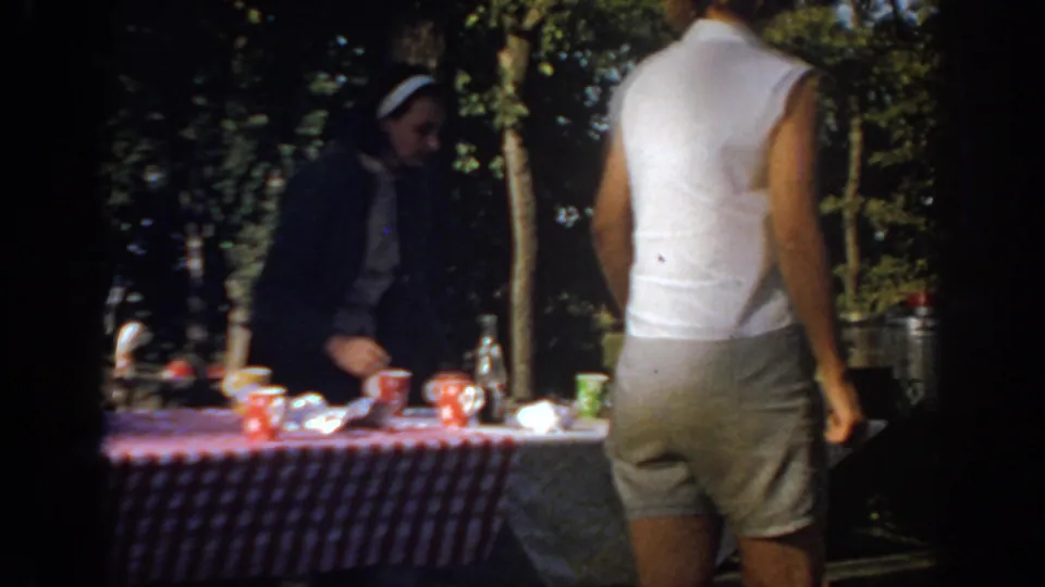 1967 Oklahoma home-movie frame of a woman at a picnic table with cups and bottles as another adult stands close by.